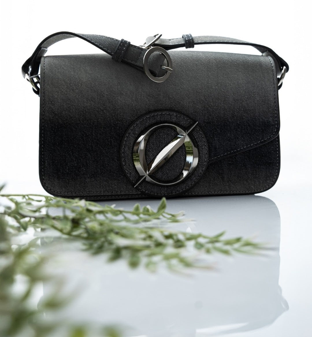 Black handbag with a metallic logo on a reflective surface with greenery