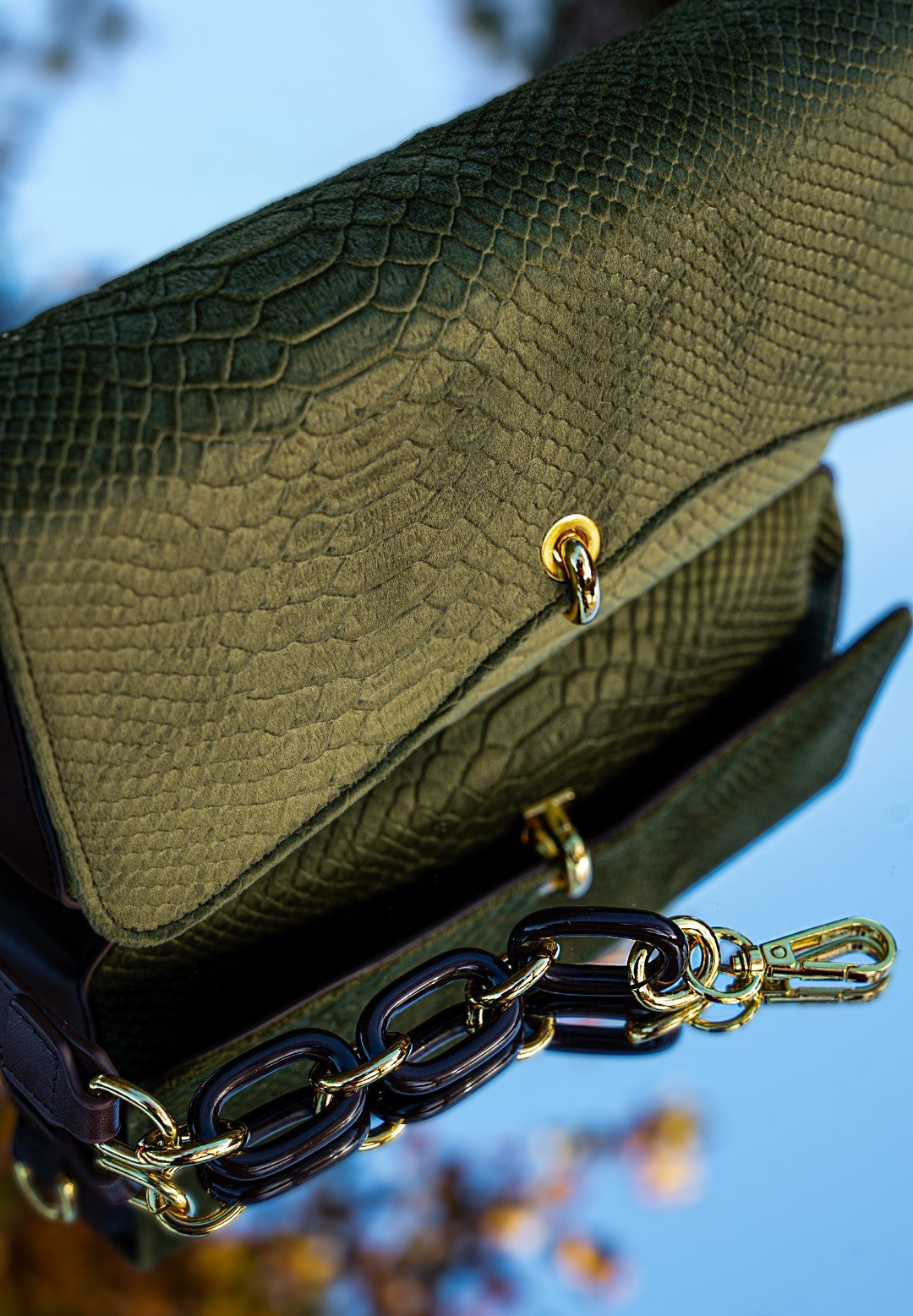 Green textured handbag with gold chain detail against a blurred natural background