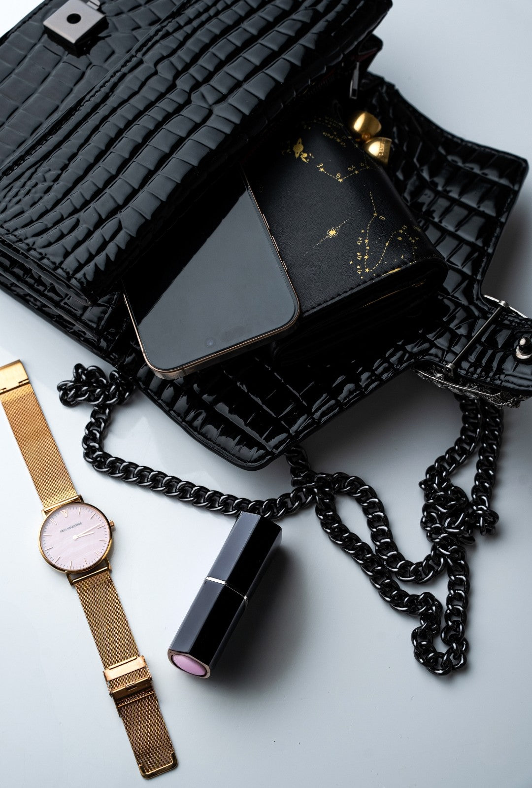 Black textured handbag with a phone, watch, and lipstick on a light gray background