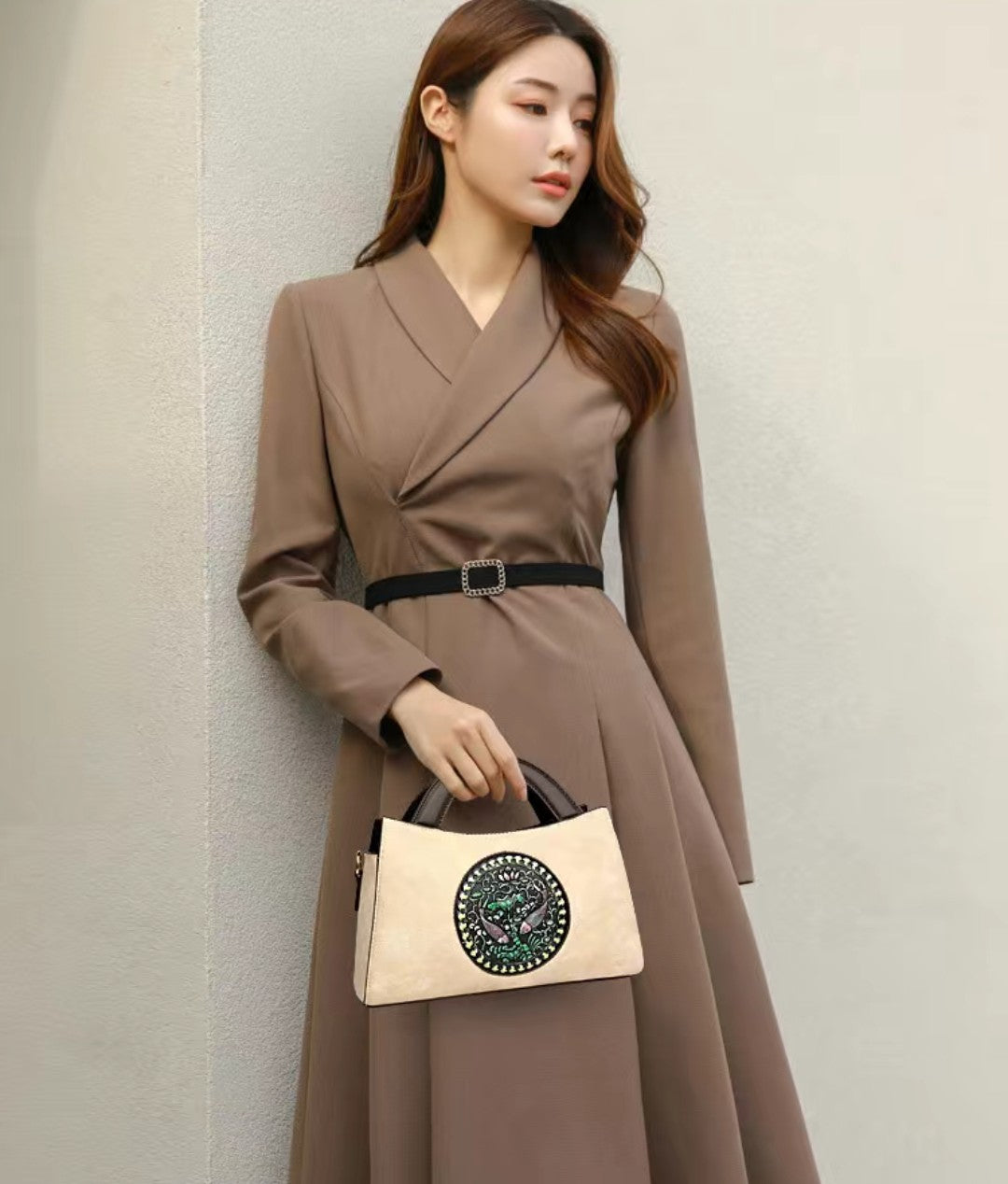 Woman in a brown dress holding a beige handbag against a plain background