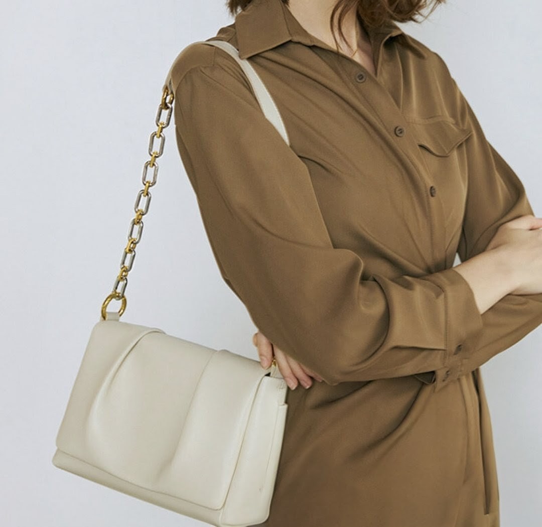 Person wearing a brown coat holding a beige handbag with a gold chain strap on a light gray background