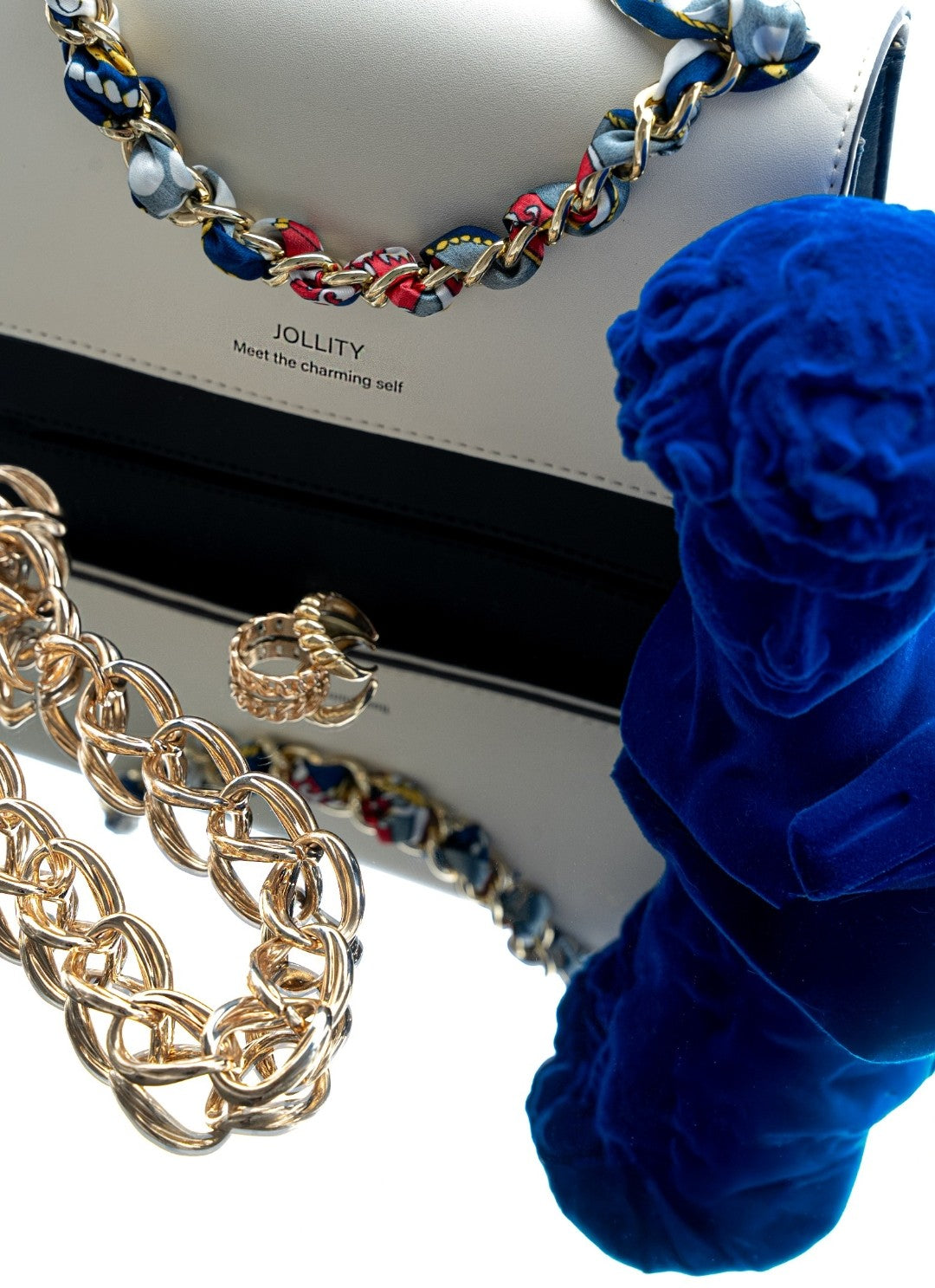 Gold chain necklace, blue flower, and Jollity handbag on a white background
