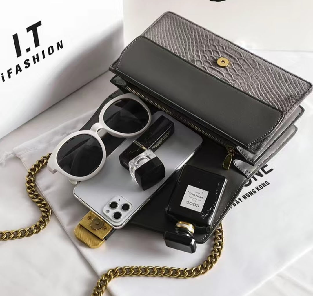Black handbag with gold chain, sunglasses, phone case, and perfume bottle on a white surface with 'I.T Fashion' branding.