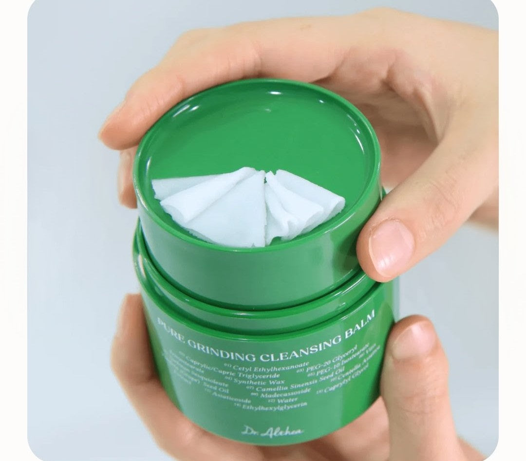 Green jar of cleansing balm with cotton pads held by a hand on a light gray background