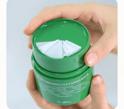 Green jar of cleansing balm with cotton pads held by a hand on a light gray background