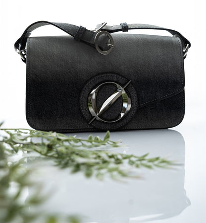 Black handbag with a metallic logo on a reflective surface with greenery