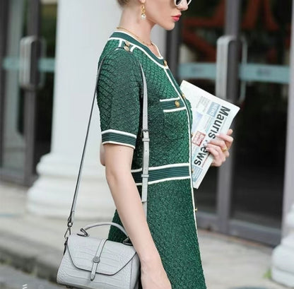 Woman in a green dress holding a newspaper and a white handbag outdoors.
