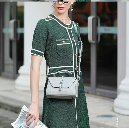 Woman in a green dress holding a gray bag and a newspaper, standing in front of a building entrance.