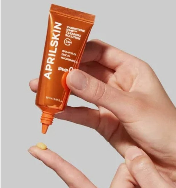 Hand holding an orange skincare product tube with 'AprilSkin' branding against a light gray background