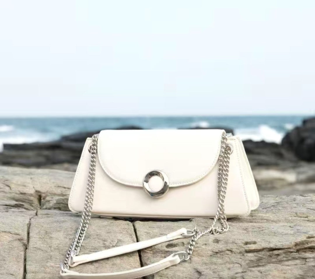 White handbag with a chain detail on a wooden surface near the ocean.
