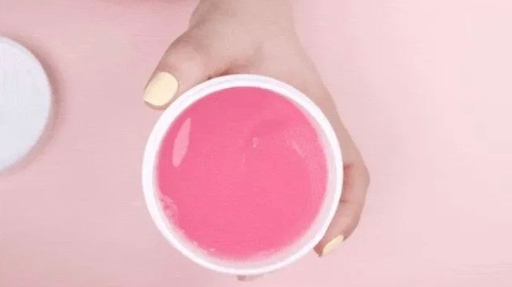 Hand holding a small white container with pink substance against a pink background